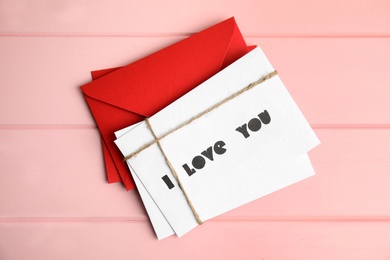 Card with text I Love You and red envelopes on pink wooden table, flat lay