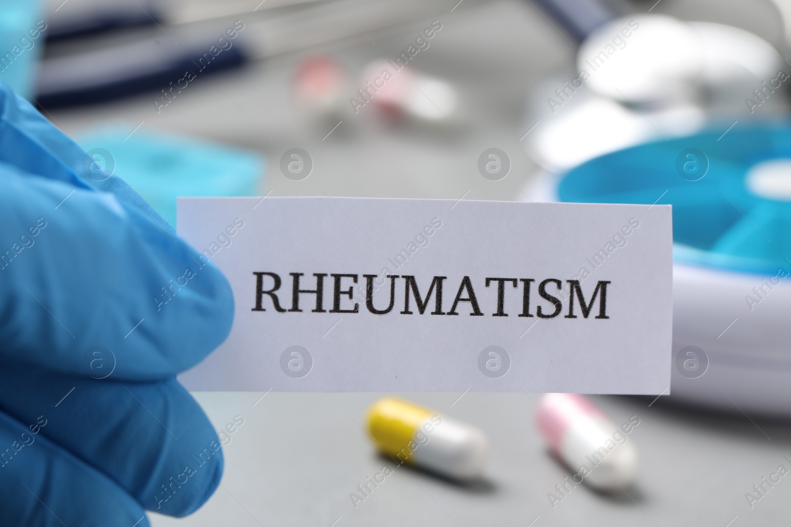 Photo of Doctor in glove holding sheet of paper with word Rheumatism above table with pills, closeup