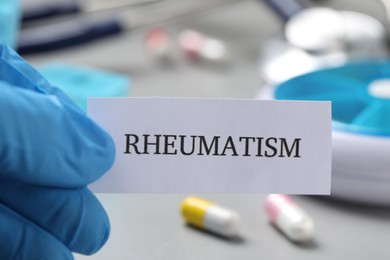 Photo of Doctor in glove holding sheet of paper with word Rheumatism above table with pills, closeup