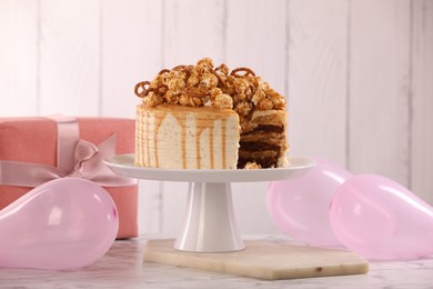 Photo of Caramel drip cake decorated with popcorn and pretzels near balloons and gift on white marble table
