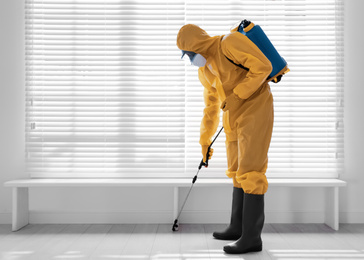 Photo of Male worker in protective suit spraying insecticide on wooden floor indoors, space for text. Pest control