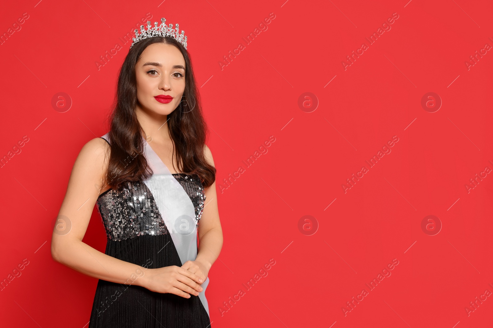Photo of Beautiful young woman with tiara and ribbon in dress on red background, space for text. Beauty contest