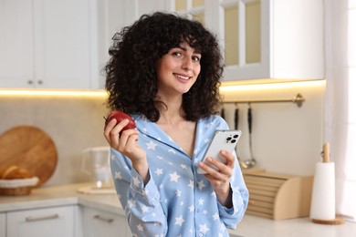 Photo of Beautiful young woman in stylish pyjama with smartphone and fresh apple in kitchen