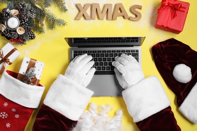Photo of Santa Claus using laptop, closeup. Gift boxes and Christmas decor on yellow background, top view