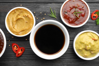 Photo of Different tasty sauces in bowls, parsley, chili pepper and rosemary on black wooden table, flat lay