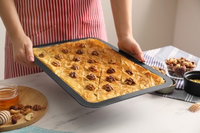 Photo of Woman holding baking pan of delicious baklava with walnuts above white marble table, closeup