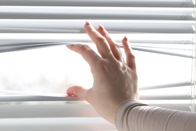 Photo of Woman separating slats of white blinds indoors, closeup