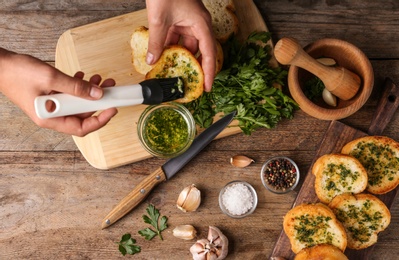 Photo of Woman brushing slice of toasted bread with garlic and herbs over wooden table, top view