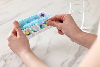 Woman with pills, organizer and glass of water at white marble table, closeup