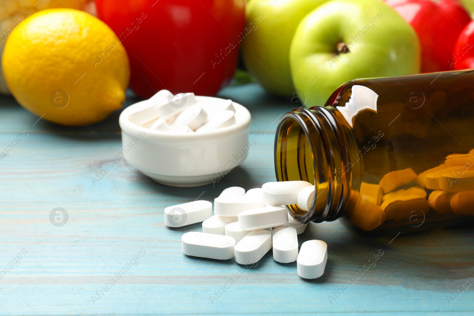 Photo of Dietary supplements. Overturned bottle and bowl with pills near food products on light blue wooden table, closeup