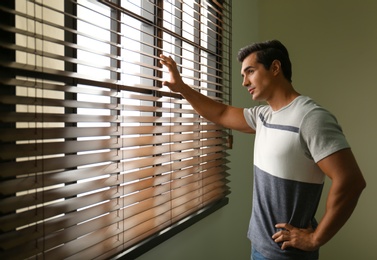 Handsome young man looking through window blinds indoors