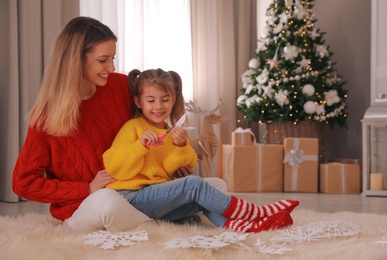 Happy mother and daughter making paper snowflake near Christmas tree at home