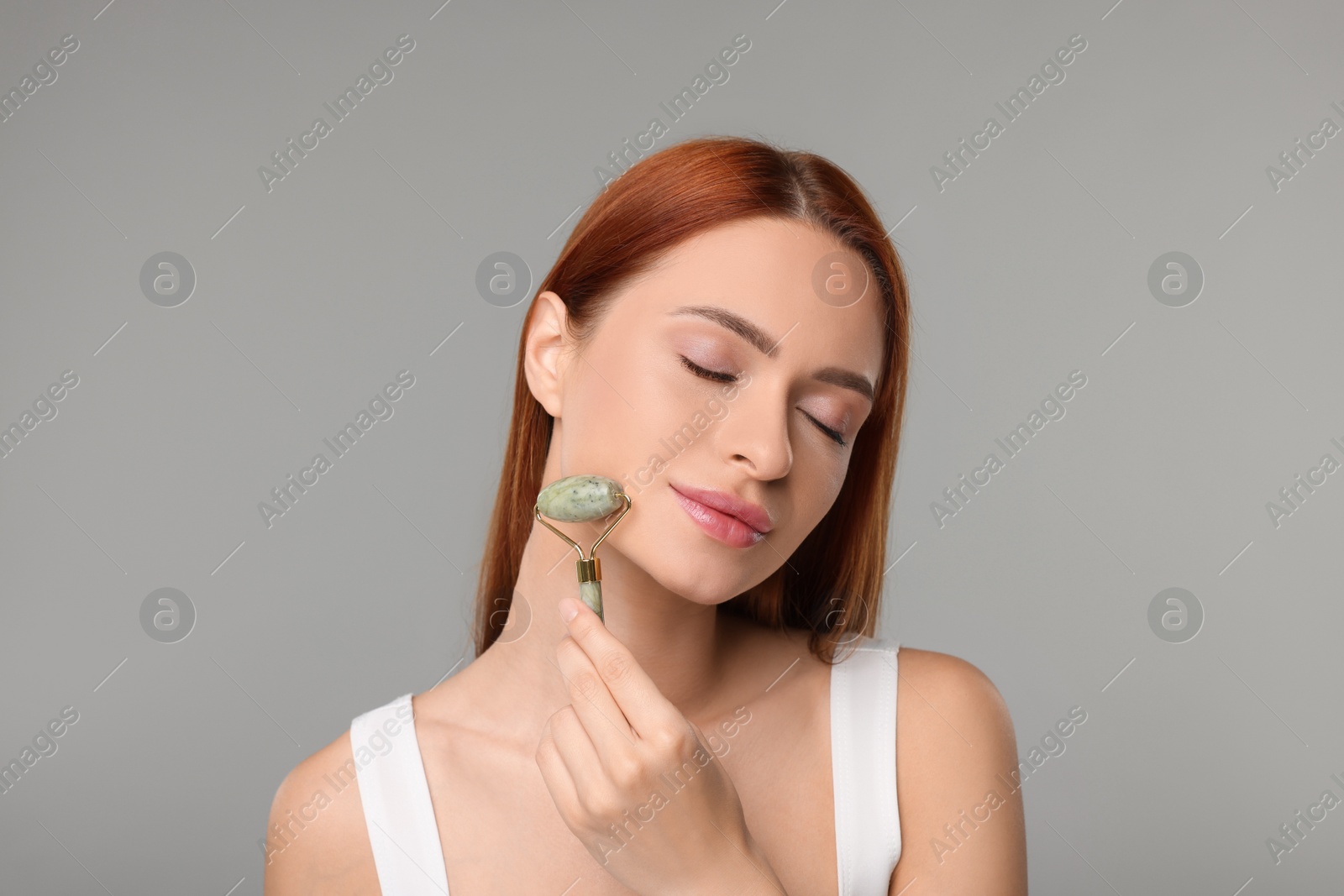 Photo of Young woman massaging her face with jade roller on grey background