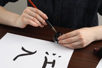 Photo of Calligraphy. Woman with brush and inkwell writing hieroglyphs on paper at wooden table, closeup