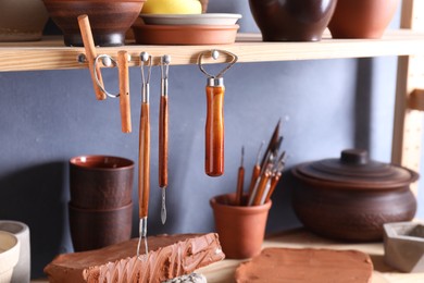 Photo of Set of different crafting tools and clay dishes on wooden rack in workshop, closeup