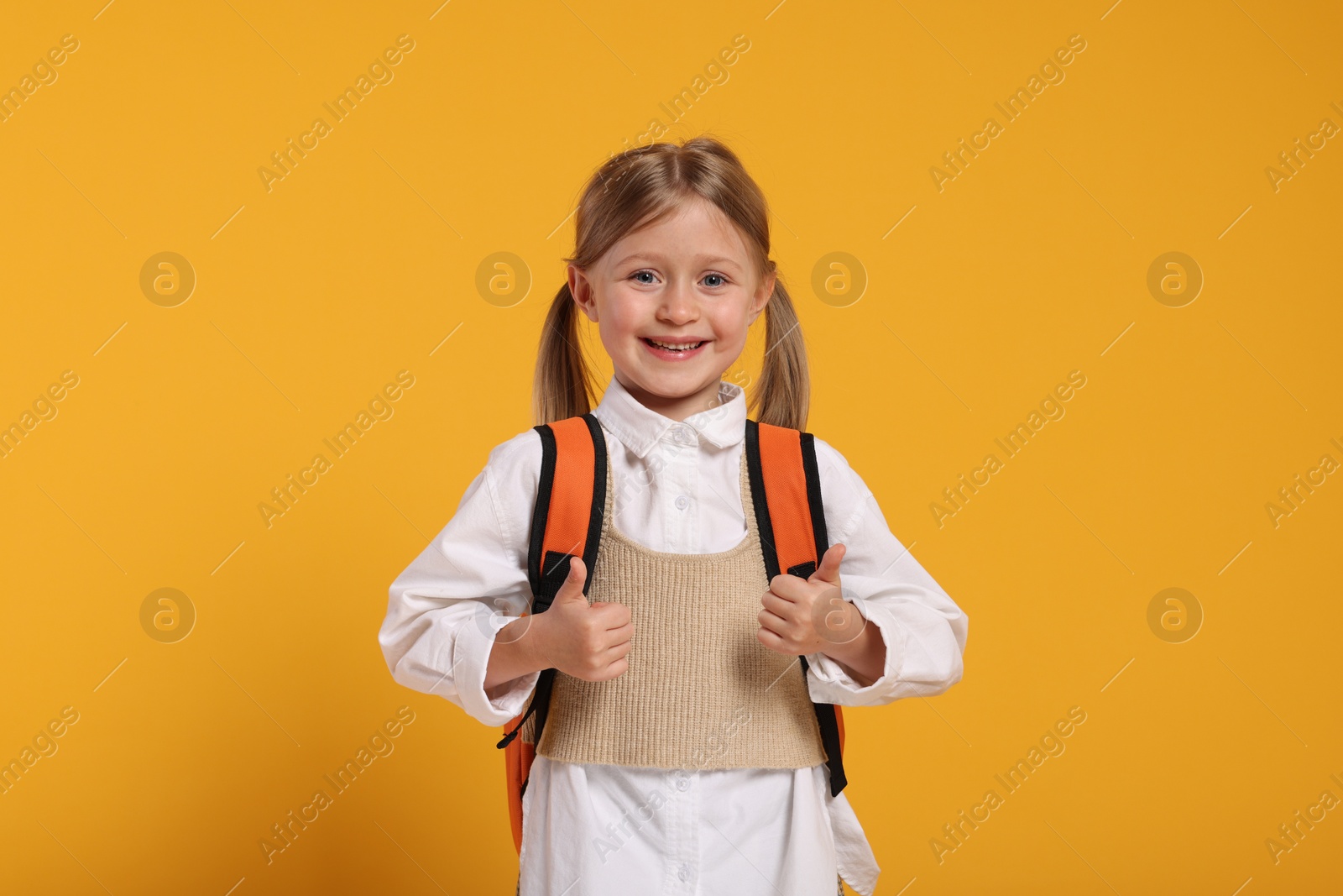 Photo of Happy schoolgirl with backpack showing thumbs up gesture on orange background