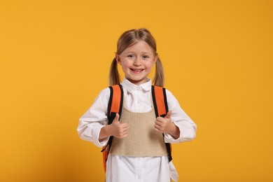 Happy schoolgirl with backpack showing thumbs up gesture on orange background