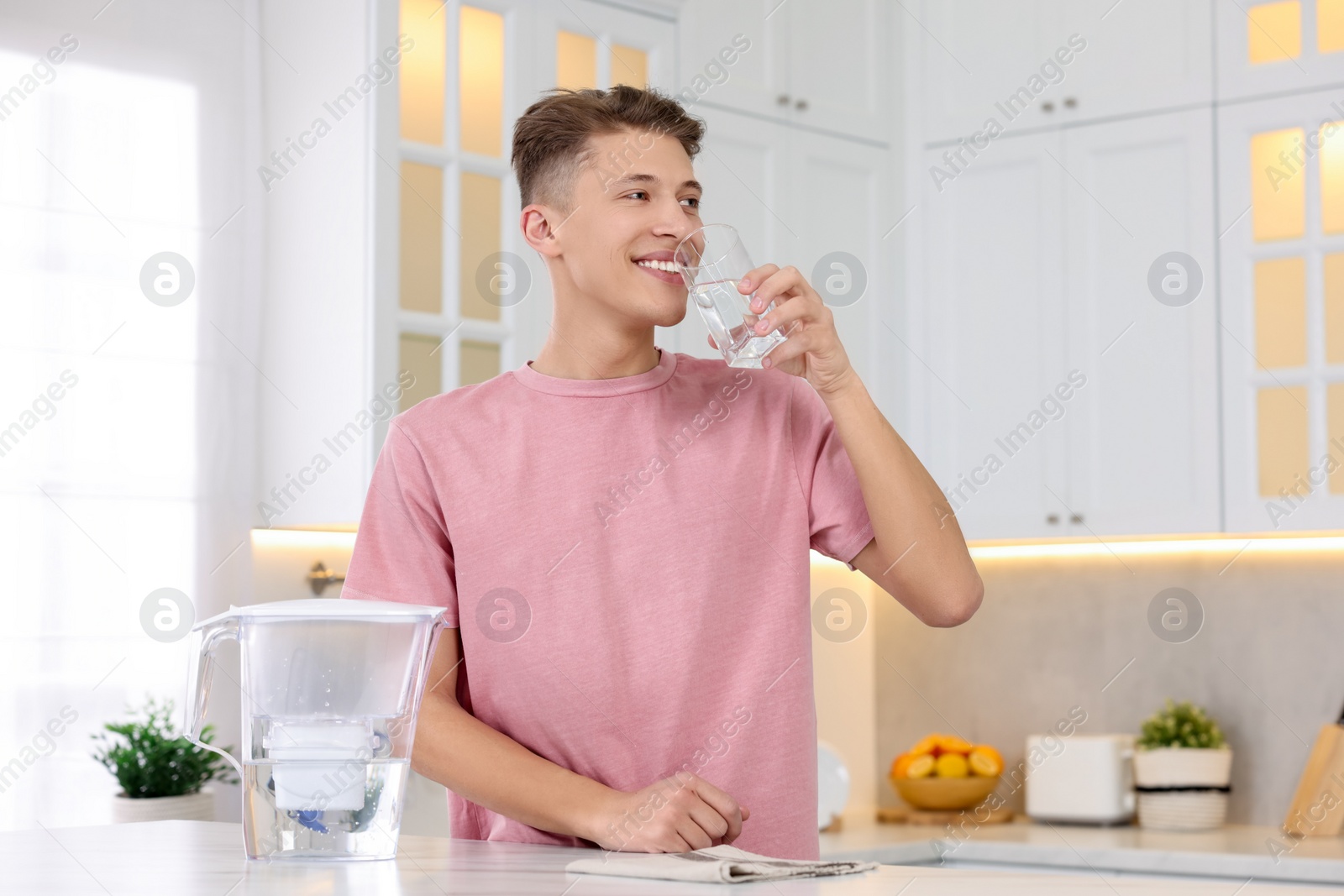 Photo of Happy man drinking clear water near filter jug at table in kitchen