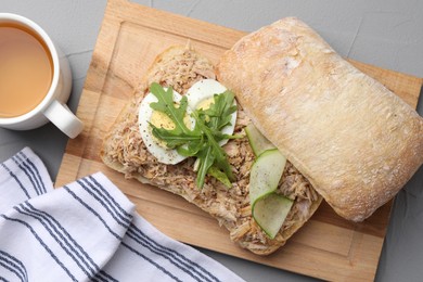 Delicious sandwich with tuna, boiled egg, vegetables and cup of tea on light grey table, flat lay