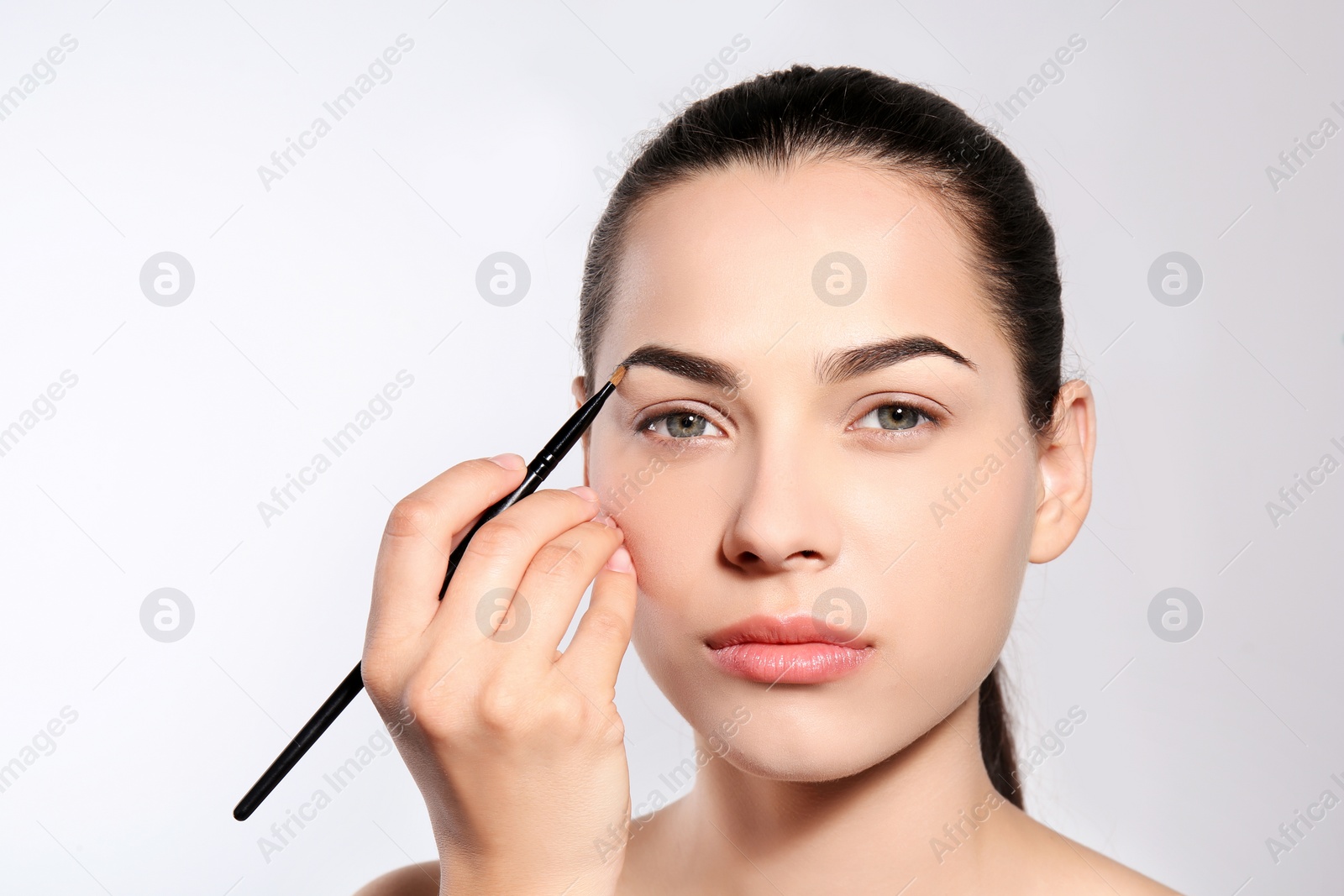 Photo of Young woman correcting shape of eyebrow with brush on light background