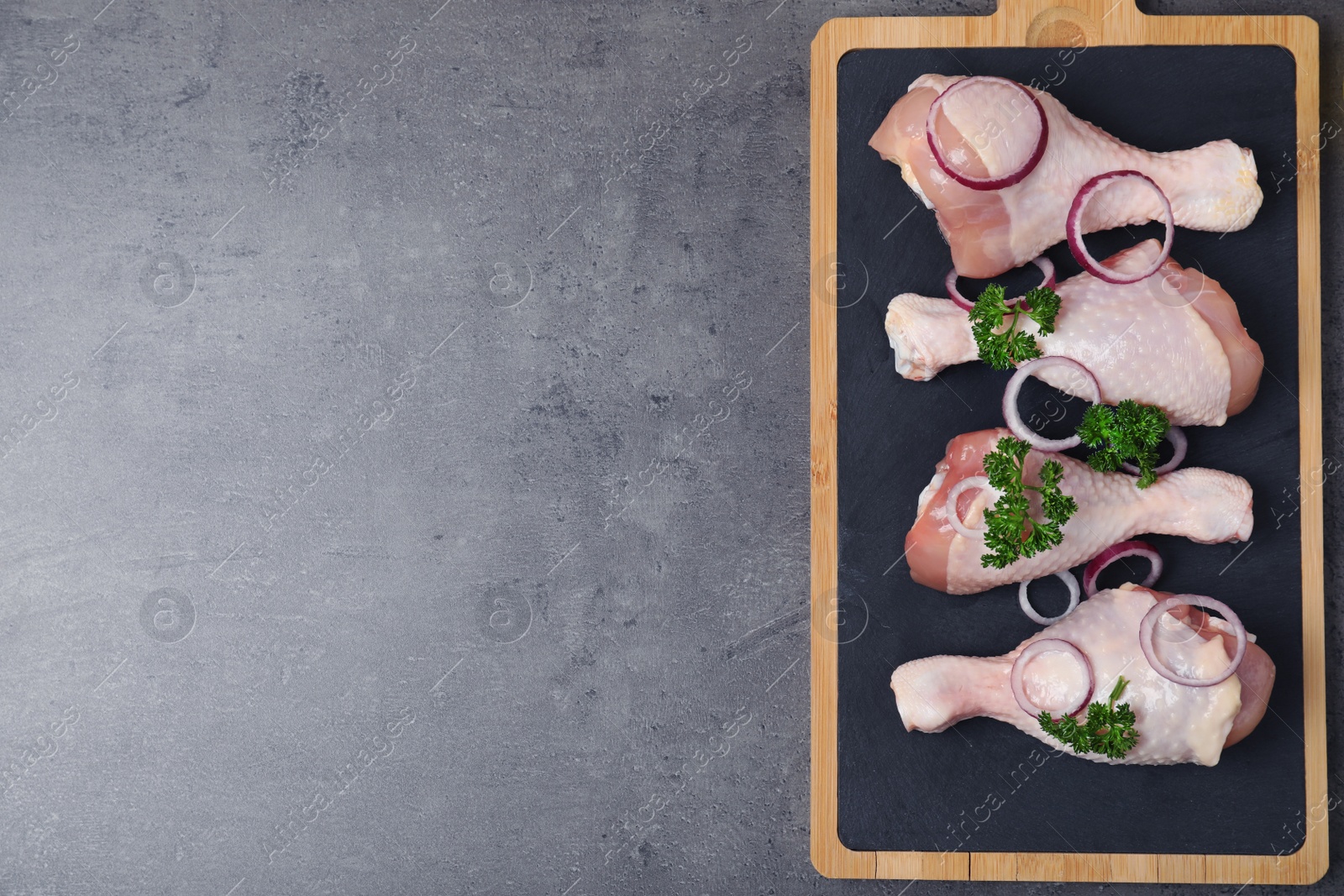 Photo of Board with raw chicken meat, onion rings and parsley on grey background, top view. Space for text