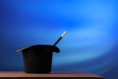 Photo of Magician's hat and wand on wooden table against blue background, space for text