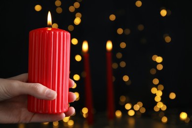 Woman holding burning red candle against blurred Christmas lights, closeup. Space for text