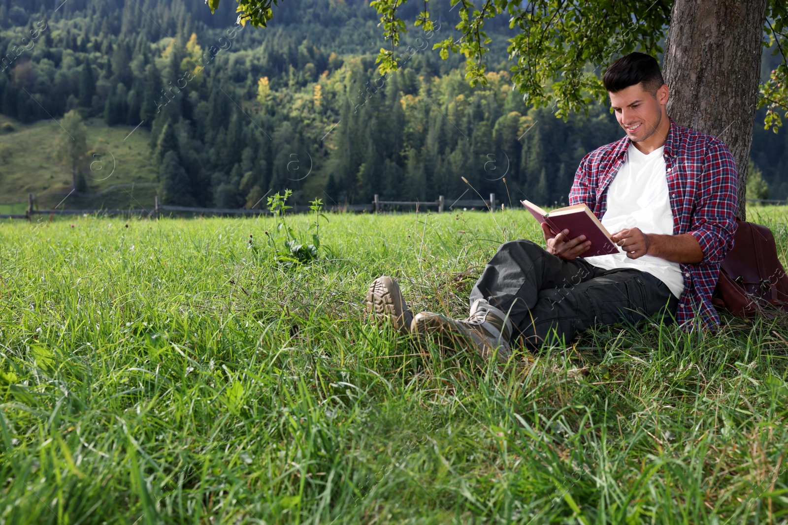 Photo of Handsome man reading book under tree on green meadow near forest