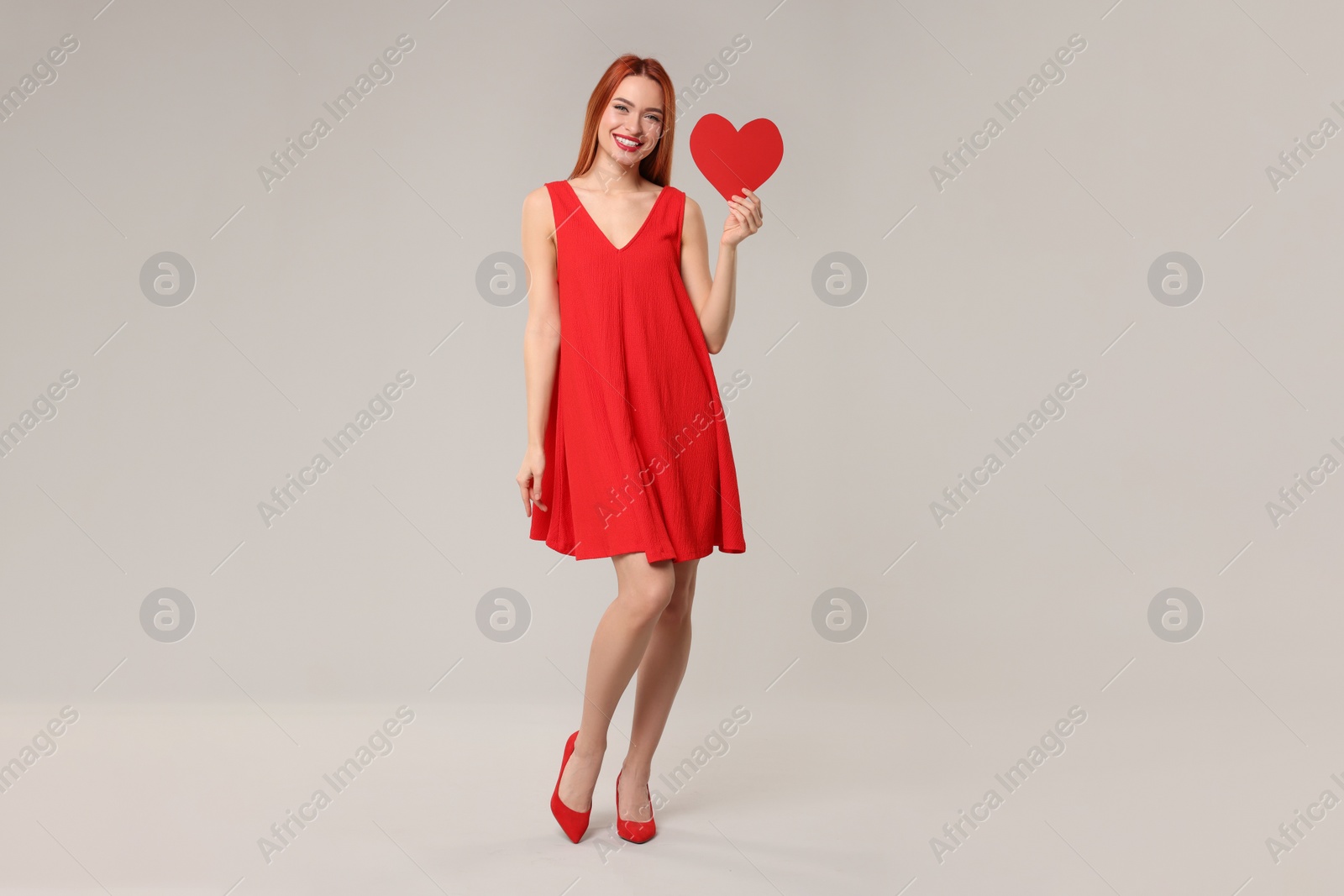 Photo of Young woman in red dress with paper heart on light grey background