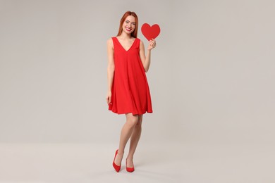 Photo of Young woman in red dress with paper heart on light grey background
