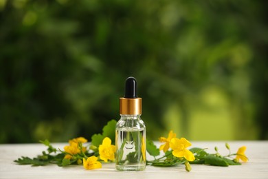 Photo of Bottle of natural celandine oil near flowers on white wooden table outdoors