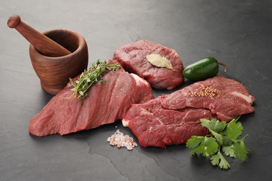 Pieces of raw beef meat, mortar with pestle and spices on black table