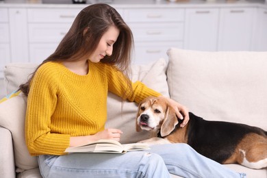Photo of Happy young woman reading book near her cute Beagle dog on couch at home. Lovely pet
