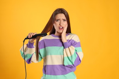 Photo of Stressed young woman with flattening iron on yellow background. Hair damage