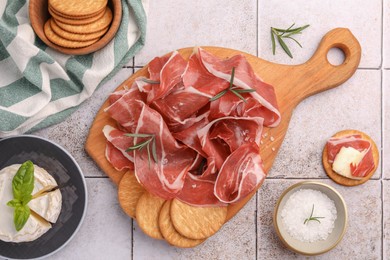 Slices of tasty cured ham, rosemary and crackers on tiled table, flat lay