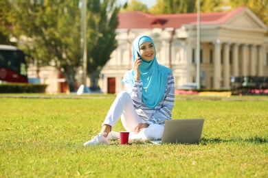 Photo of Muslim woman in hijab with laptop talking on phone outdoors