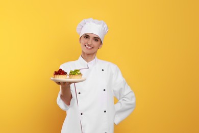 Happy professional confectioner in uniform with delicious tartlets on yellow background