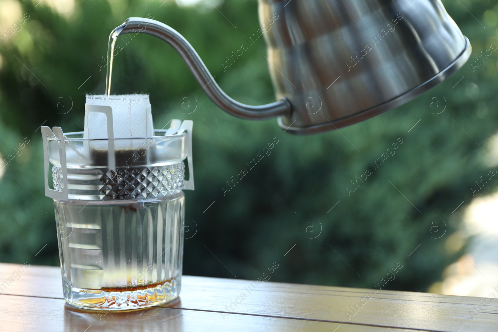 Photo of Pouring hot water into glass with drip coffee bag from kettle on wooden table, closeup