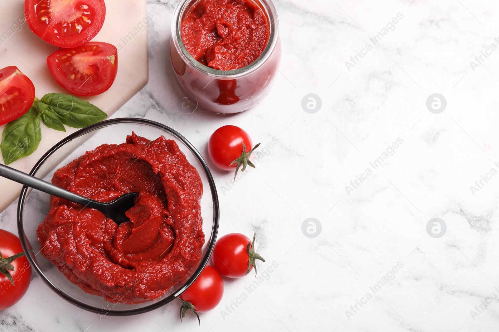 Photo of Glass bowl and jar of tasty tomato paste with spoon and ingredients on white marble table, flat lay. Space for text