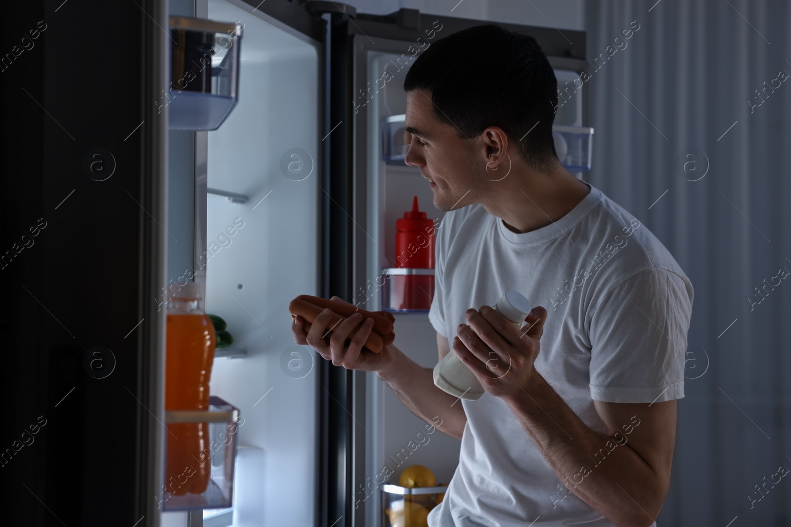 Photo of Happy man with sausages and sauce near refrigerator in kitchen at night