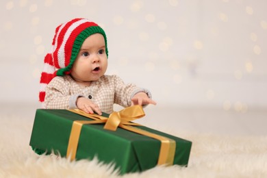 Photo of Cute baby in elf hat with Christmas gift on fluffy carpet against blurred festive lights, space for text. Winter holiday