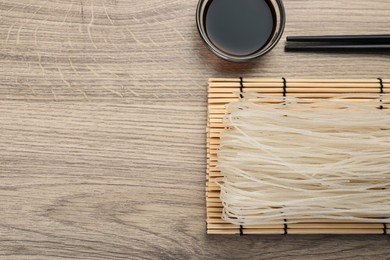 Photo of Dried rice noodles with chopsticks and soy sauce on wooden table, flat lay. Space for text