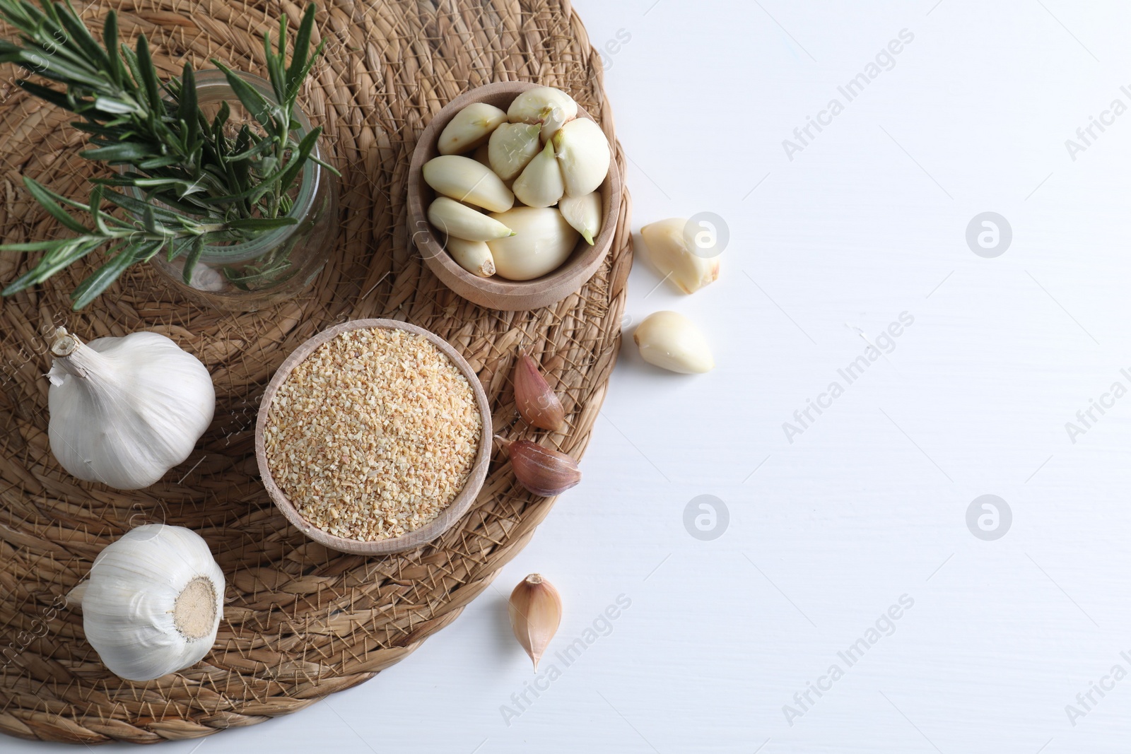 Photo of Dehydrated garlic granules in bowl, rosemary and fresh cloves on white table, flat lay. Space for text