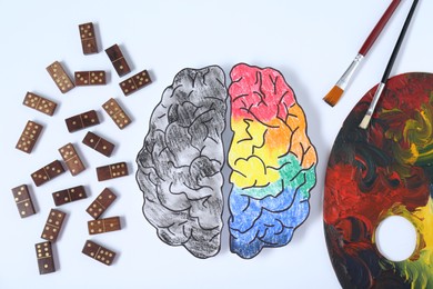 Photo of Logic and creativity. Paper brain with one colorful part and another grey on white background, flat lay. Domino tiles near left hemisphere and palette with brushes near right one