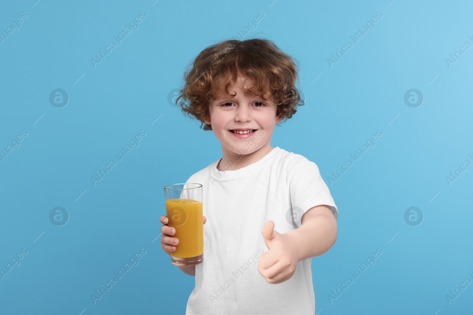 Photo of Cute little boy with glass of fresh juice showing thumb up on light blue background