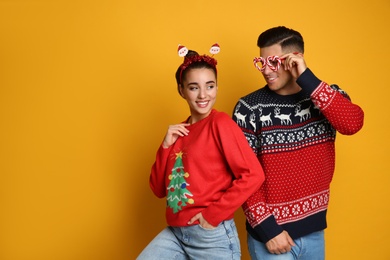Couple in Christmas sweaters, Santa headband and party glasses on yellow background, space for text