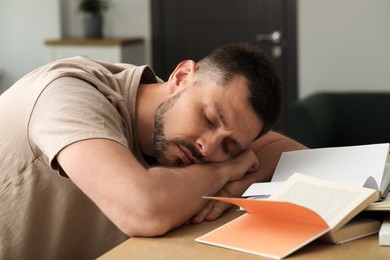 Photo of Tired man sleeping among books at wooden table indoors