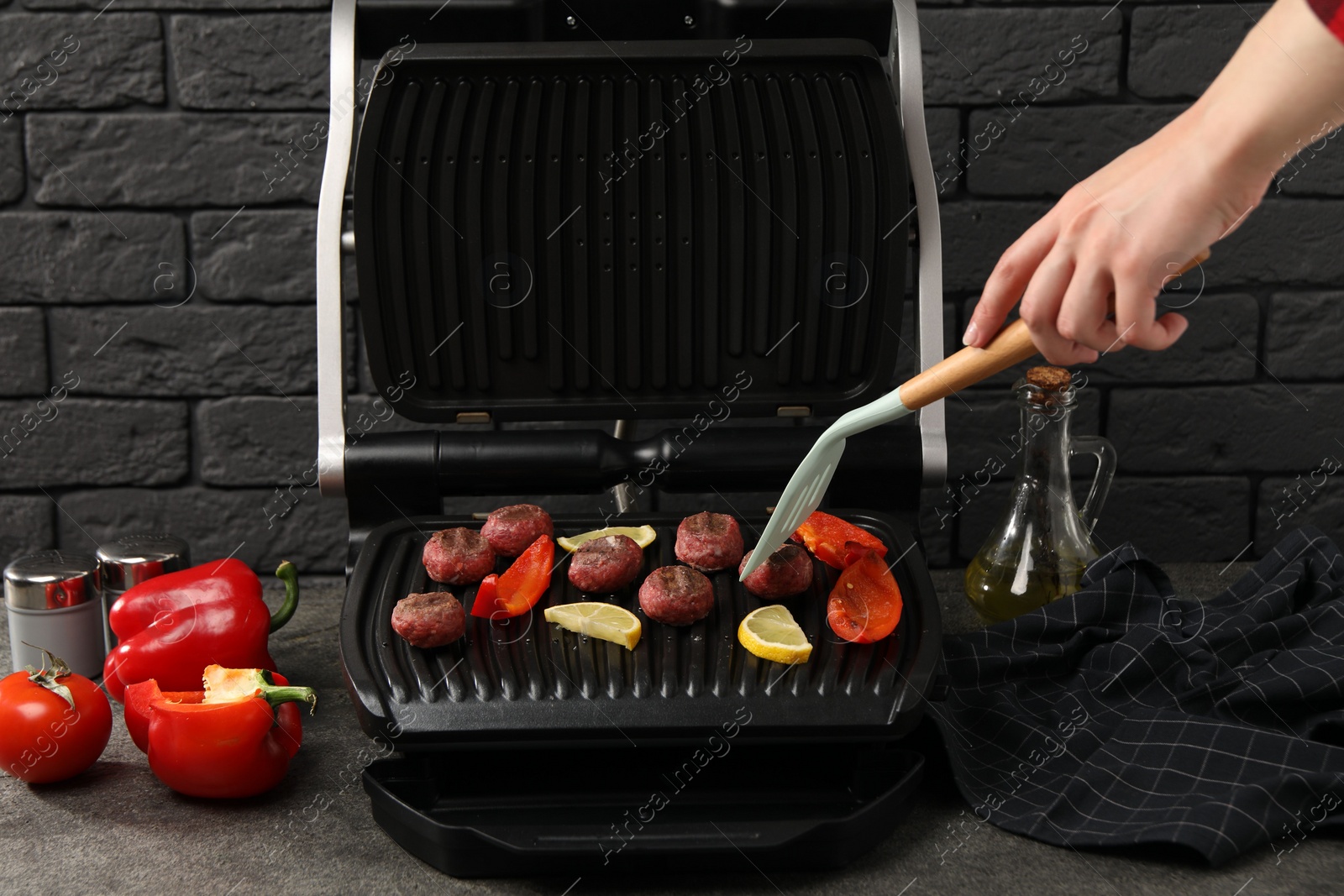 Photo of Woman cooking meat balls with bell peppers and lemon on electric grill at grey textured table, closeup