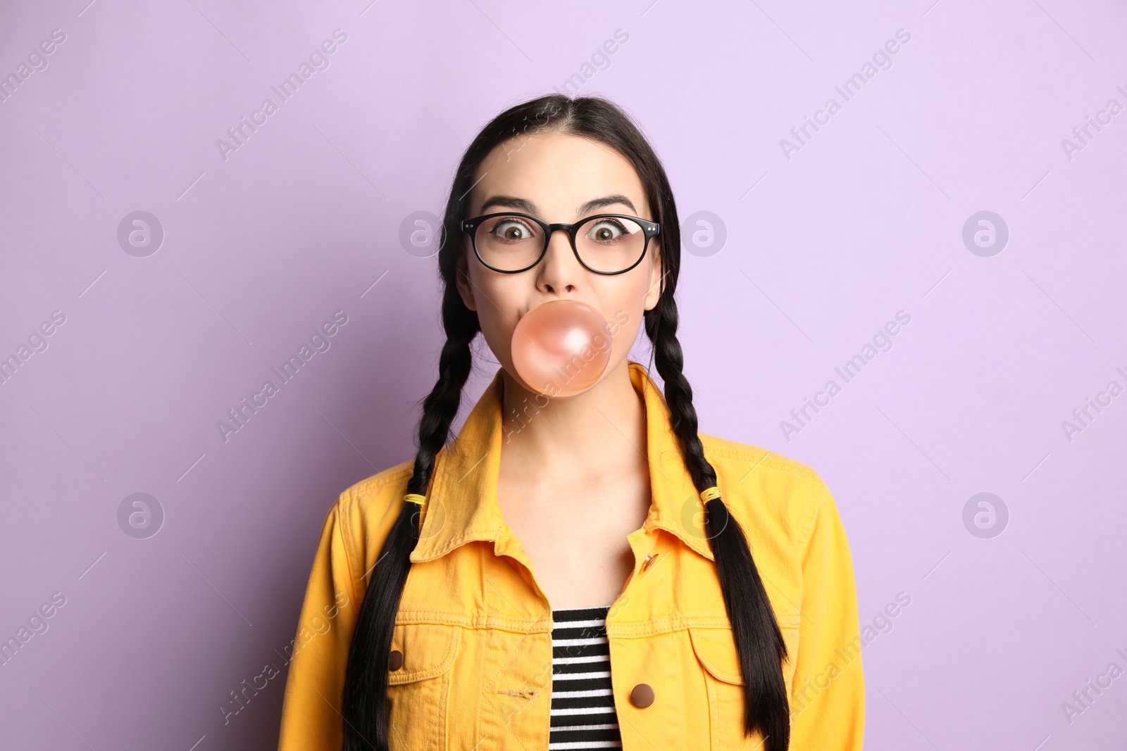 Photo of Fashionable young woman with braids blowing bubblegum on lilac background