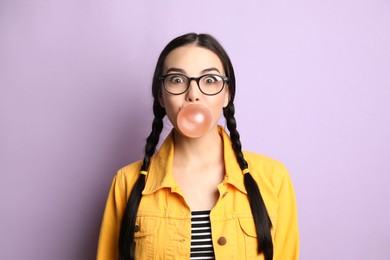 Photo of Fashionable young woman with braids blowing bubblegum on lilac background